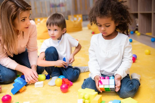 Educador y niños multiétnicos jugando con constructor de plástico en el jardín de infantes - foto de stock