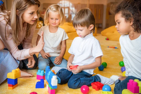 Multicultural kids playing with constructor, educator gesturing in kindergarten — Stock Photo