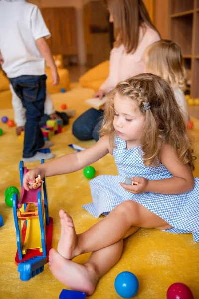 Entzückendes Kind im Kleid sitzt auf Teppich und spielt im Kindergarten mit Spielzeug — Stockfoto