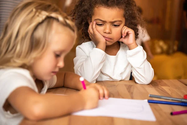 Enfant afro-américain insatisfait regardant dessin enfant caucasien à table à la maternelle — Photo de stock