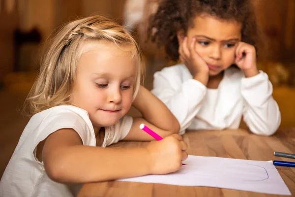 Niño afroamericano aburrido mirando hacia otro lado, niño caucásico dibujo en la mesa en el jardín de infantes - foto de stock