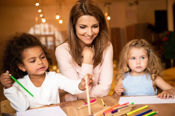 Educatore allegro seduto vicino a bambini multiculturali disegno nella scuola materna — Foto stock