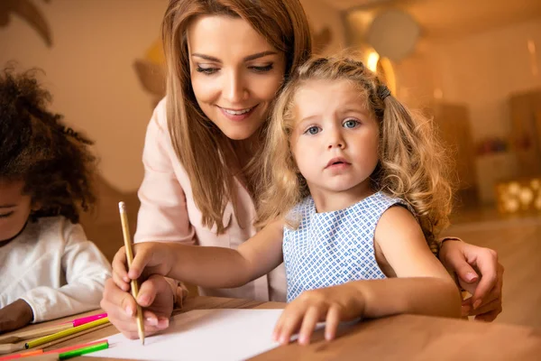 Educador alegre ayudando a niño adorable dibujo en el jardín de infantes - foto de stock