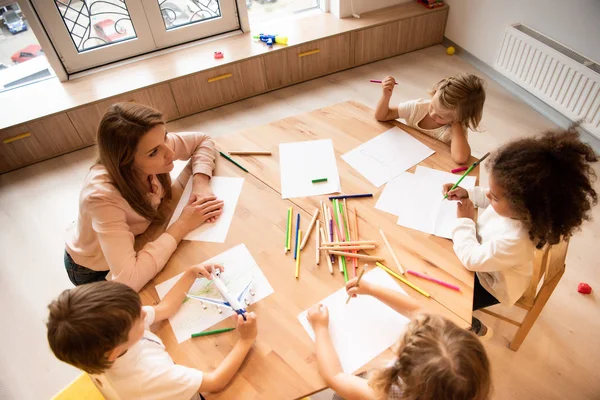 Vista de ángulo alto del educador multicultural y los niños dibujando juntos en la mesa en el jardín de infantes - foto de stock