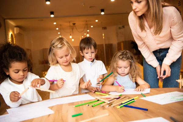 Educatore guardando gruppo multietnico di bambini che disegnano nella scuola materna — Foto stock