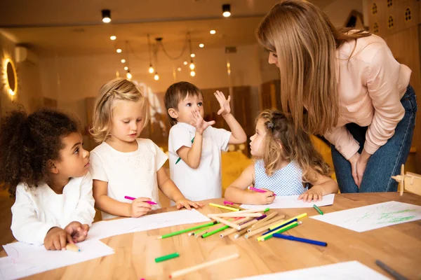 Enfants multiethniques dessinant à la maternelle, éducateur regardant le garçon gestuel — Photo de stock