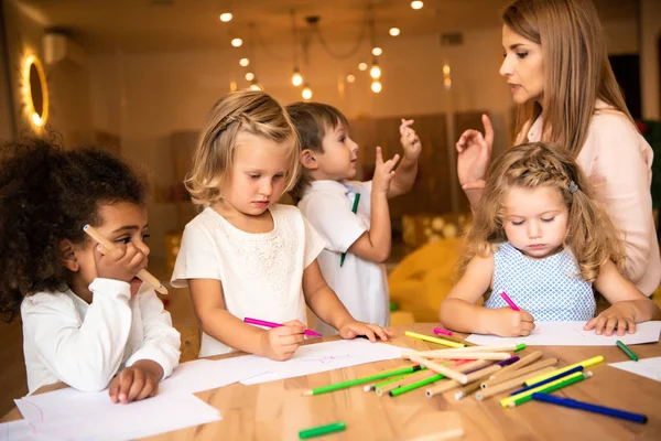 Multicultural kids drawing in kindergarten, educator looking at gesturing boy — Stock Photo