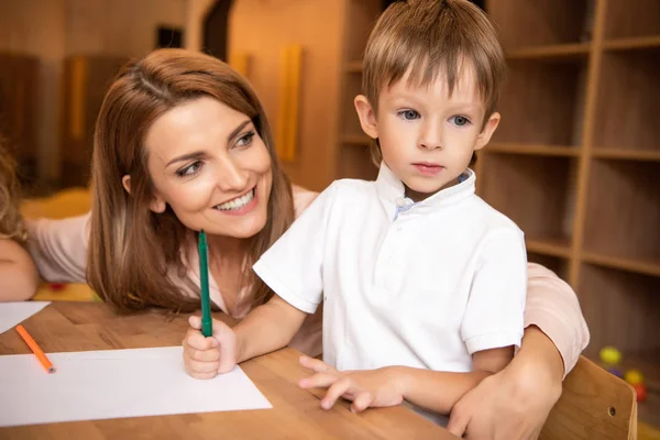 Educatore sorridente abbracciare adorabile ragazzo a tavola nella scuola materna — Foto stock