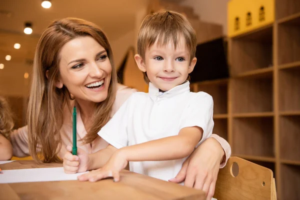 Счастливый педагог обнимает очаровательного мальчика за столом в детском саду — стоковое фото