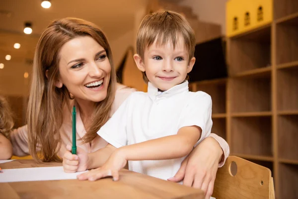 Feliz educador abrazando adorable niño en la mesa en el jardín de infantes - foto de stock