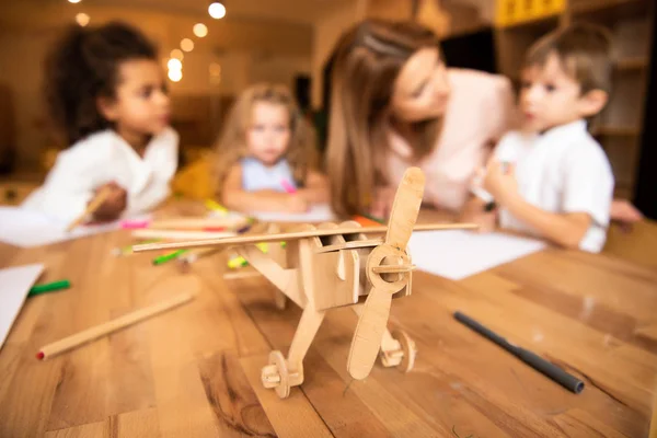 Педагог и мультиэтнические дети рисуют в детском саду, деревянный самолет на переднем плане — стоковое фото