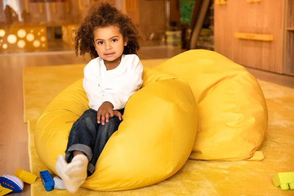 Adorable enfant afro-américain assis sur une chaise de sac de haricot à la maternelle et regardant la caméra — Photo de stock