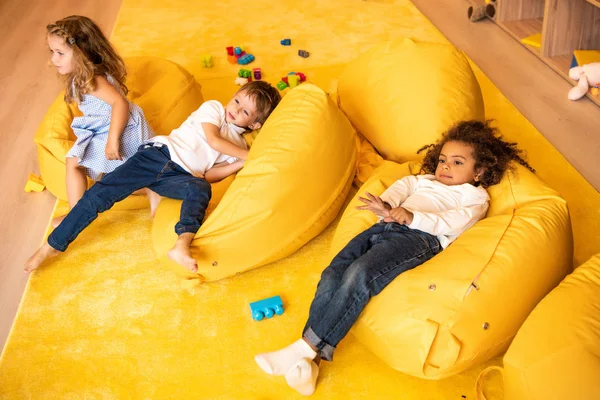 High angle view of multicultural children lying on yellow bean bag chairs in kindergarten — Stock Photo