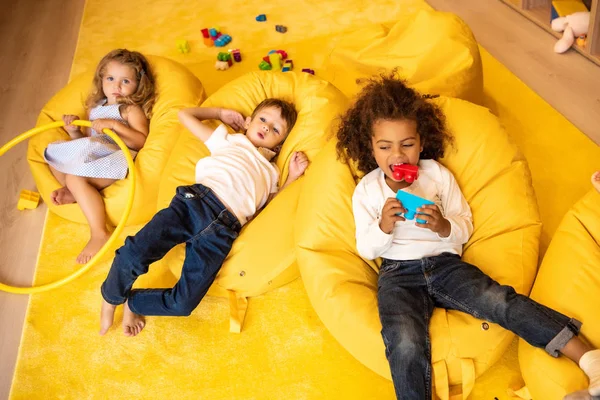 Visão de alto ângulo de crianças multiétnicas deitadas em cadeiras de saco de feijão com brinquedos no jardim de infância — Fotografia de Stock