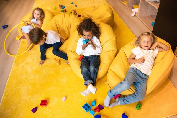 Vista de ángulo alto de los niños multiculturales tumbados en sillas de bolsa de frijol en el jardín de infantes — Stock Photo