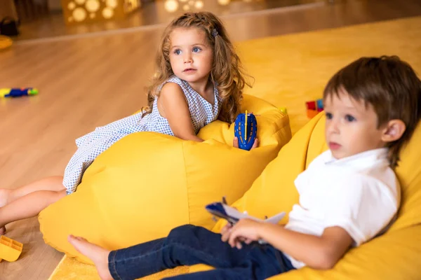 Adorables niños con juguetes tumbados en sillas de bolsa de frijol amarillo en el jardín de infantes y mirando hacia otro lado - foto de stock