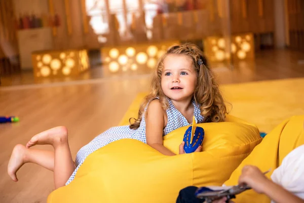 Sonriente adorable niño acostado en la silla bolsa de frijol con juguete y mirando hacia otro lado en el jardín de infantes - foto de stock