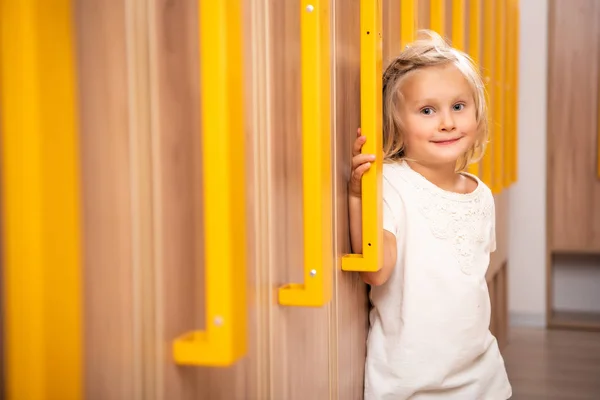 Fröhliches liebenswertes Kind, das in der Garderobe des Kindergartens steht und in die Kamera schaut — Stockfoto