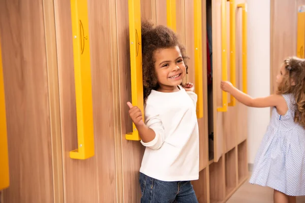 Happy multicultural kids standing near lockers in kindergarten cloakroom — Stock Photo