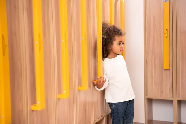 Adorable enfant afro-américain debout près des casiers en bois dans le vestiaire de la maternelle — Photo de stock