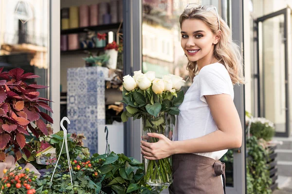 Attraente fiorista in possesso di vaso con rose bianche vicino negozio di fiori e guardando la fotocamera — Foto stock
