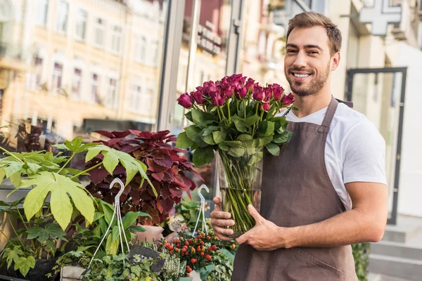 Красивый улыбающийся флорист, держащий розы в банке рядом с цветочным магазином — стоковое фото