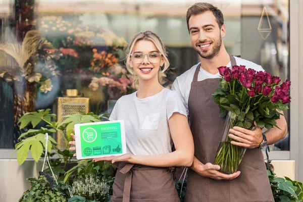 Floristas felizes segurando tablet com aparelho de compras e rosas borgonhas perto de loja de flores — Fotografia de Stock