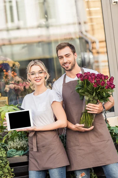 Fleuristes souriants tenant tablette avec écran blanc et roses bordeaux près de la boutique de fleurs — Photo de stock