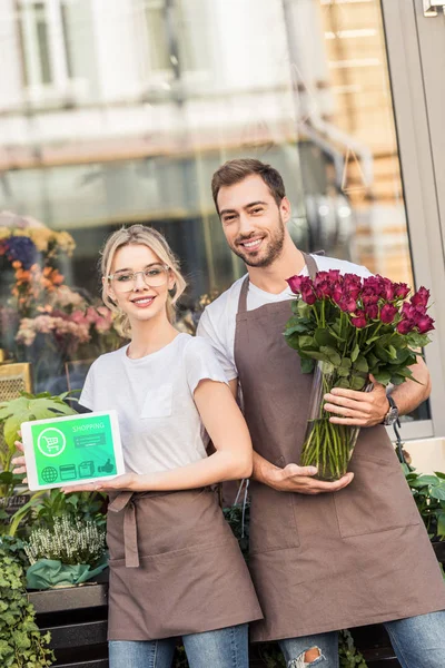 Smiling florists holding tablet with shopping appliance and burgundy roses near flower shop — Stock Photo