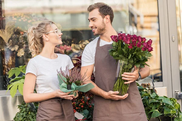 Fioristi sorridenti in piedi vicino a negozio di fiori con pianta in vaso e rose bordeaux, guardarsi l'un l'altro — Foto stock