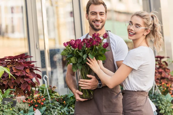 Улыбающиеся флористы, держащие бургундские розы возле цветочного магазина и смотрящие в камеру — стоковое фото