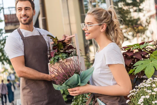 Smiling florists holding potted plants near flower shop and looking away — Stock Photo
