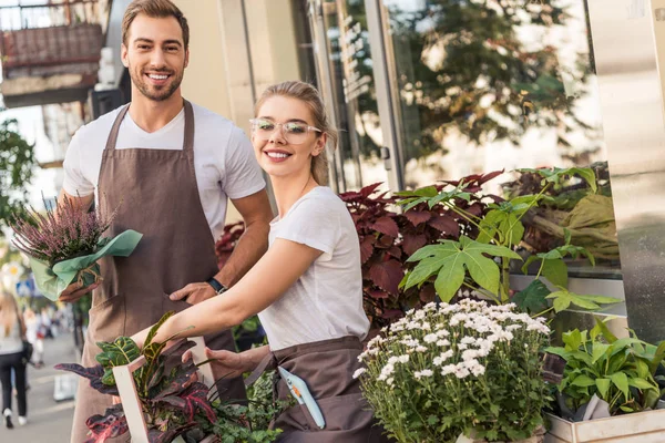 Fleuristes souriants tenant des plantes en pot près du magasin de fleurs et regardant la caméra — Photo de stock