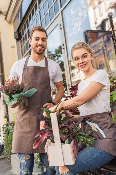Smiling colleagues holding potted plants near flower shop and looking at camera — Stock Photo
