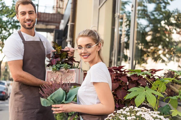 Fioristi femminili e maschili sorridenti che tengono piante in vaso vicino al negozio di fiori e guardando la fotocamera — Foto stock