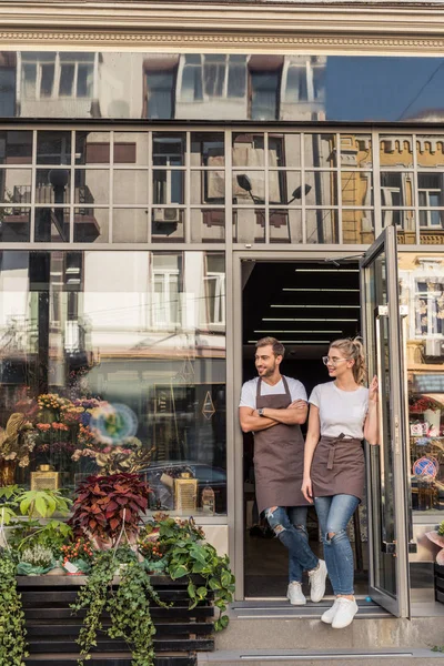 Smiling florists standing near flower shop and looking away — Stock Photo