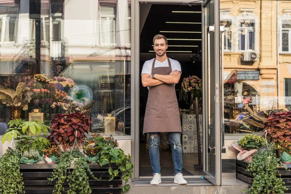 Lächelnde hübsche Floristin, die mit verschränkten Armen in der Nähe eines Blumenladens steht und in die Kamera blickt — Stockfoto