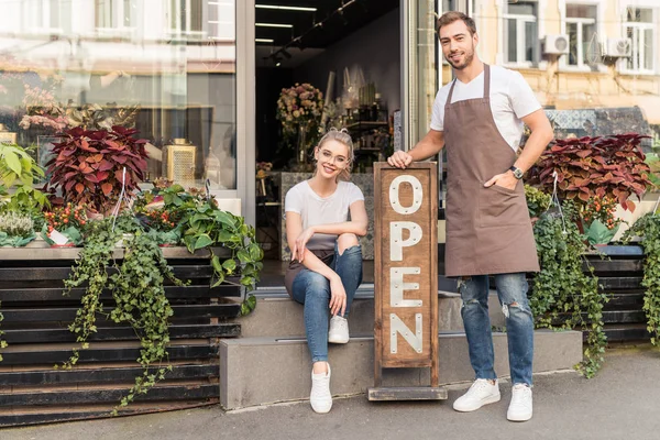 Smiling flower shop owners looking at camera on street with open signboard — Stock Photo