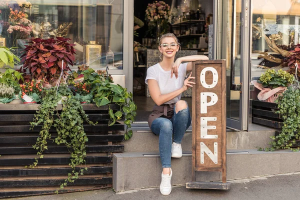 Attrayant propriétaire de magasin de fleurs souriant assis sur les escaliers avec panneau ouvert — Photo de stock