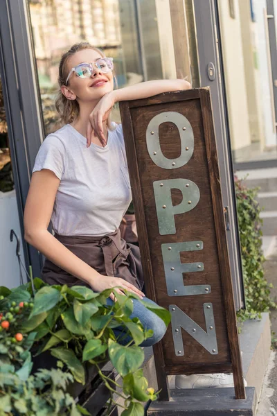 Attraente sorridente proprietario del negozio di fiori appoggiato sul segno aperto — Foto stock