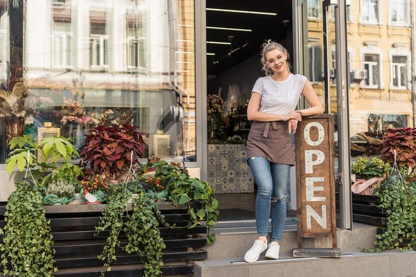 Smiling attractive florist standing on stairs of flower shop and leaning on open sign — Stock Photo