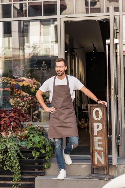 Smiling handsome florist standing on stairs of flower shop and leaning on open sign — Stock Photo