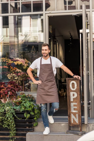 Florista guapo sonriente de pie en las escaleras de la tienda de flores y apoyado en el signo abierto - foto de stock