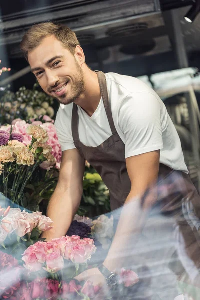 View through window of smiling handsome florist taking care of bouquets in flower shop and looking at camera — Stock Photo