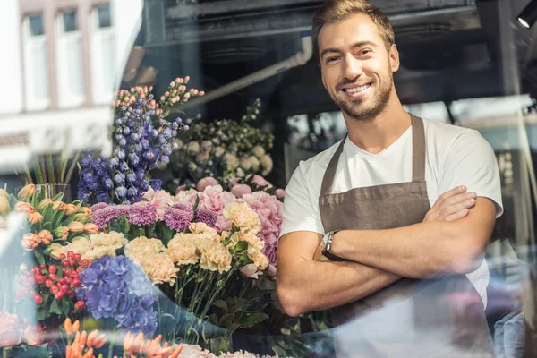 Вид через окно красивого флориста, стоящего со скрещенными руками в цветочном магазине и смотрящего в камеру — стоковое фото
