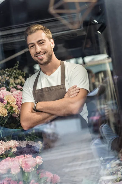 Vue à travers la fenêtre du beau fleuriste dans le tablier debout avec les bras croisés dans la boutique de fleurs et regardant la caméra — Photo de stock