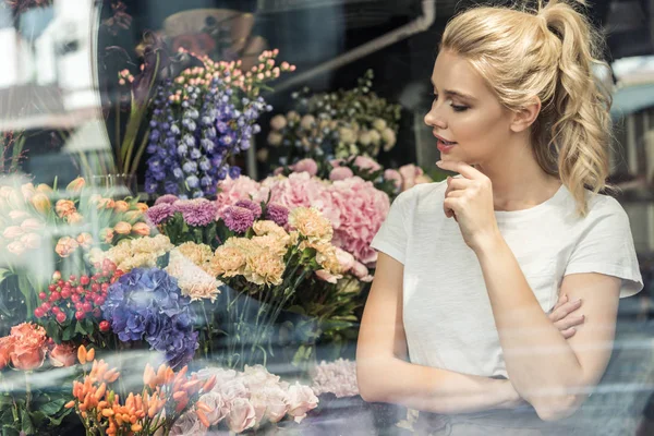 Вид через окно привлекательного задумчивого флориста, смотрящего на букеты в цветочном магазине — стоковое фото
