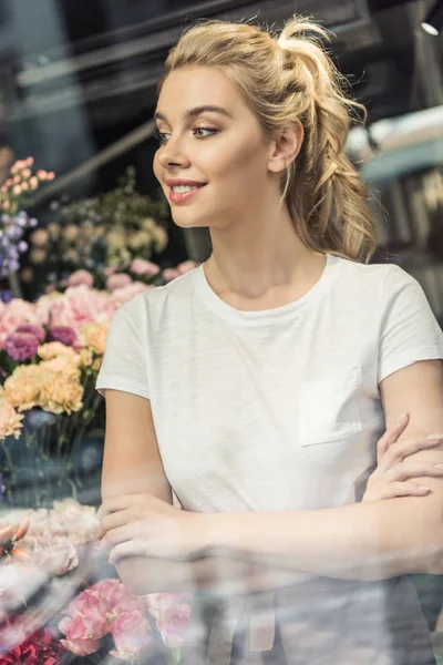 Вид через окно привлекательного флориста, стоящего со скрещенными руками в цветочном магазине и смотрящего в сторону — стоковое фото