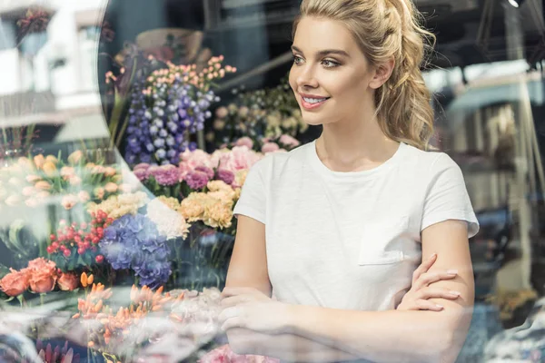 Vista a través de la ventana de la floristería hermosa de pie con brazos cruzados en la tienda de flores y mirando hacia otro lado - foto de stock