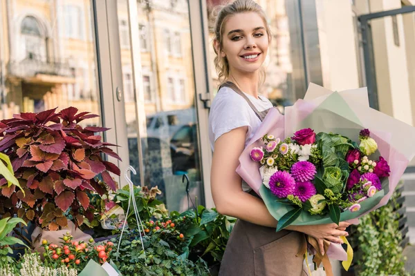 Florista sonriente sosteniendo hermoso ramo de crisantemos cerca de la tienda de flores y mirando a la cámara - foto de stock